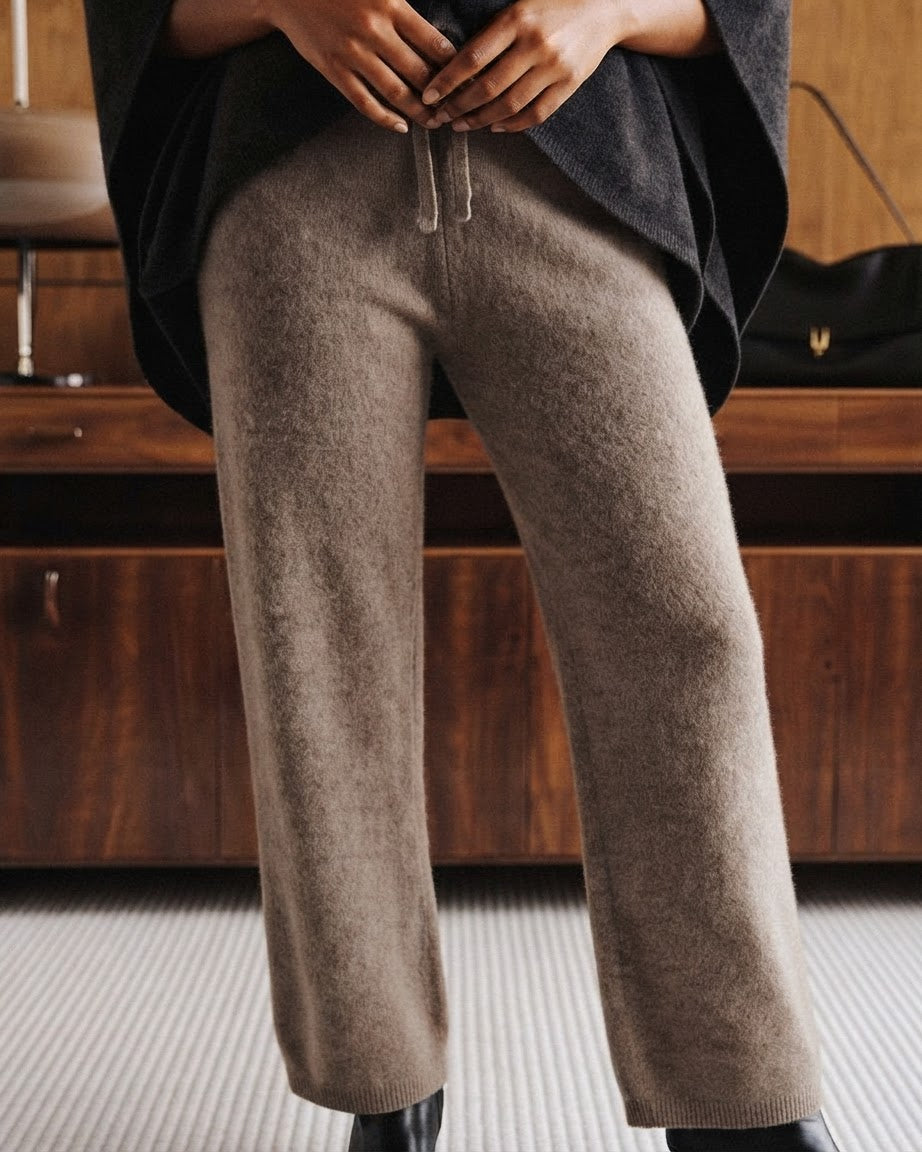 A person wearing loose, wide-leg taupe knit pants with a drawstring waist, styled with a dark charcoal top and black boots in an indoor setting with a wooden cabinet.