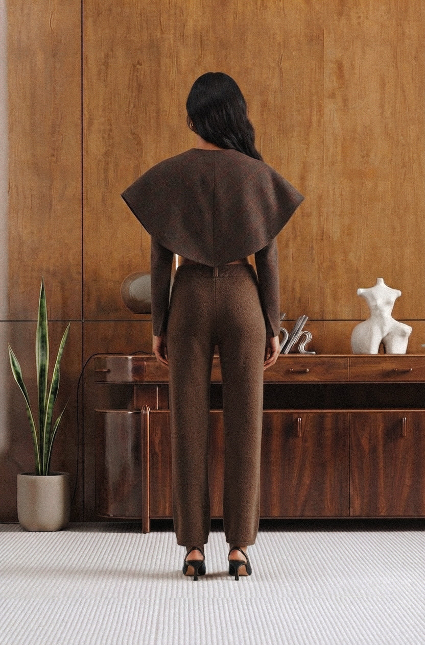 A woman stands with her back to the camera wearing a matching brown textured two-piece set consisting of a wide cape-like collar and high-waisted trousers, in a modern room with wood paneling.