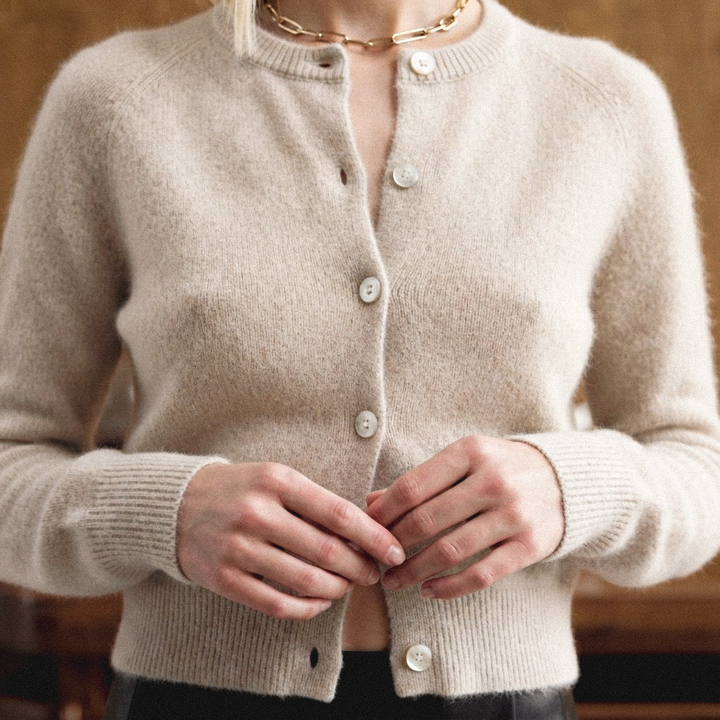 A close-up of a person wearing a soft, fuzzy beige cardigan with white buttons, shown from the neck down against a warm brown background.
