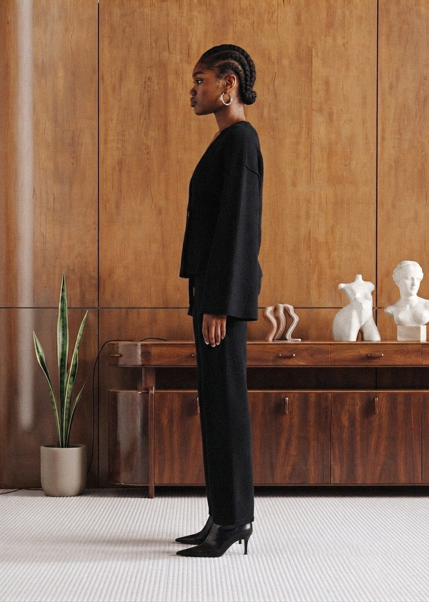 A model in a black tailored suit and black heels stands in profile in a modern interior with wood-paneled walls, a wooden console, and white sculpture busts.