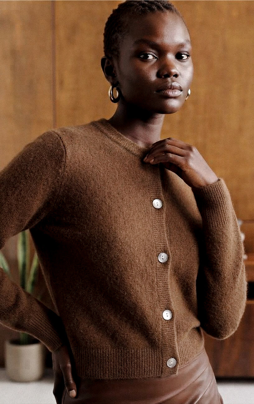 A woman wearing a brown button-front cardigan sweater, posed against a warm wood-paneled background.