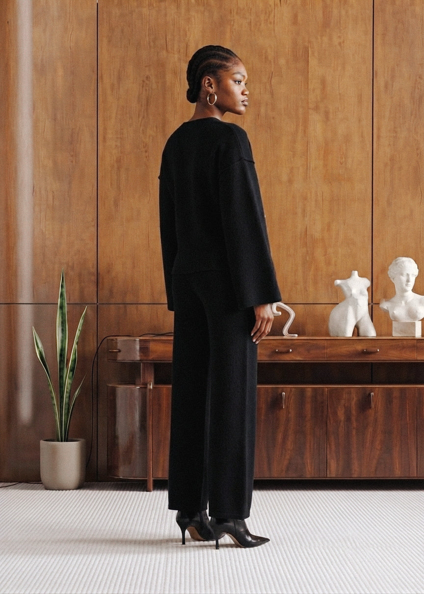 A model stands in profile wearing a black long-sleeve top and matching wide-leg trousers, posed in front of a warm wood-paneled wall with a wooden console table, white bust sculptures, and a potted plant.