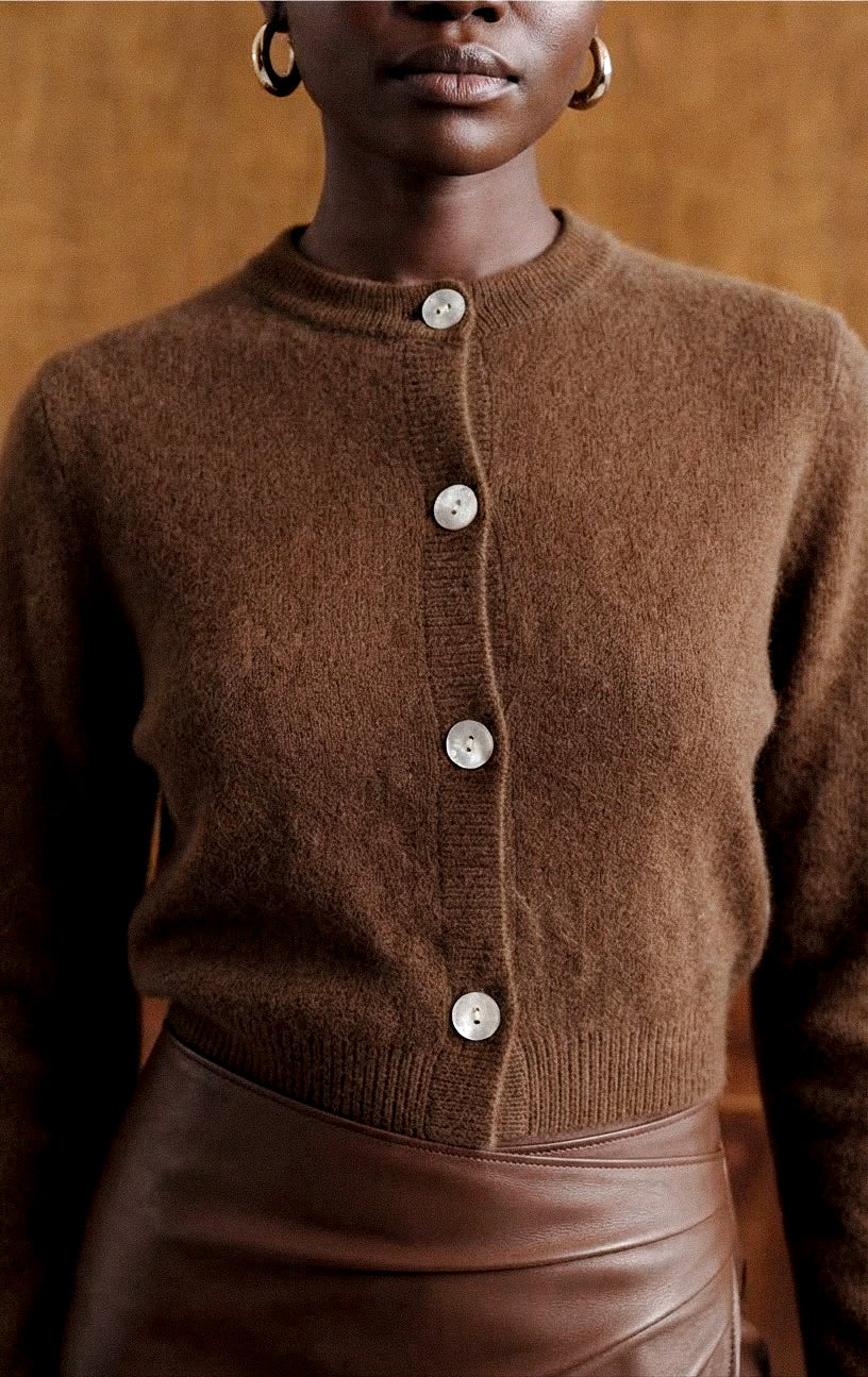 A close-up of a person wearing a brown knit cardigan with a button front and a brown leather skirt against a warm brown background.