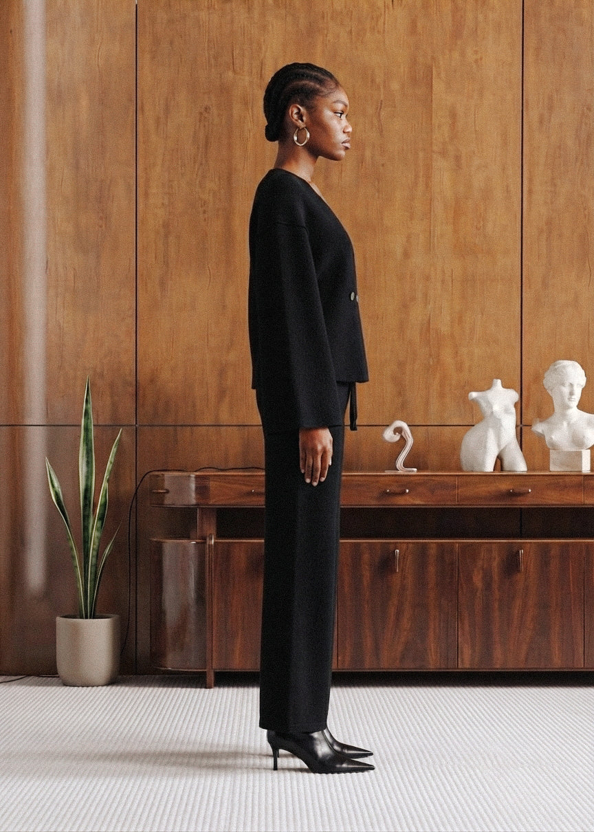 A woman stands in profile wearing a black tailored suit with a long-sleeve top and wide-leg trousers, paired with black pointed heels, in a modern interior with wood paneling and a console table.