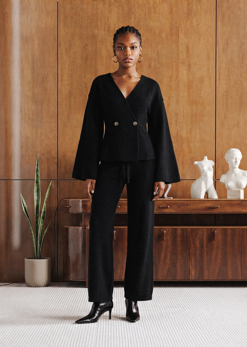 A model stands in a modern interior wearing a black double-breasted long-sleeve blazer with wide lapels and matching wide-leg trousers, paired with black pointed-toe heeled boots. The background features a warm wood-paneled wall, a wooden console table, and white decorative busts.