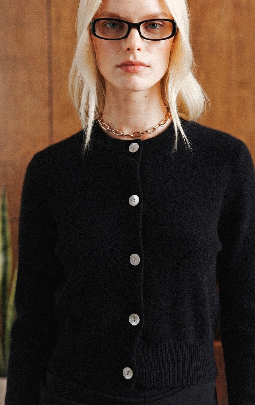 A blonde woman wearing black-rimmed glasses, a chunky gold chain necklace, and a black button-front knit cardigan sweater, shown from the chest up against a warm wood-paneled background.